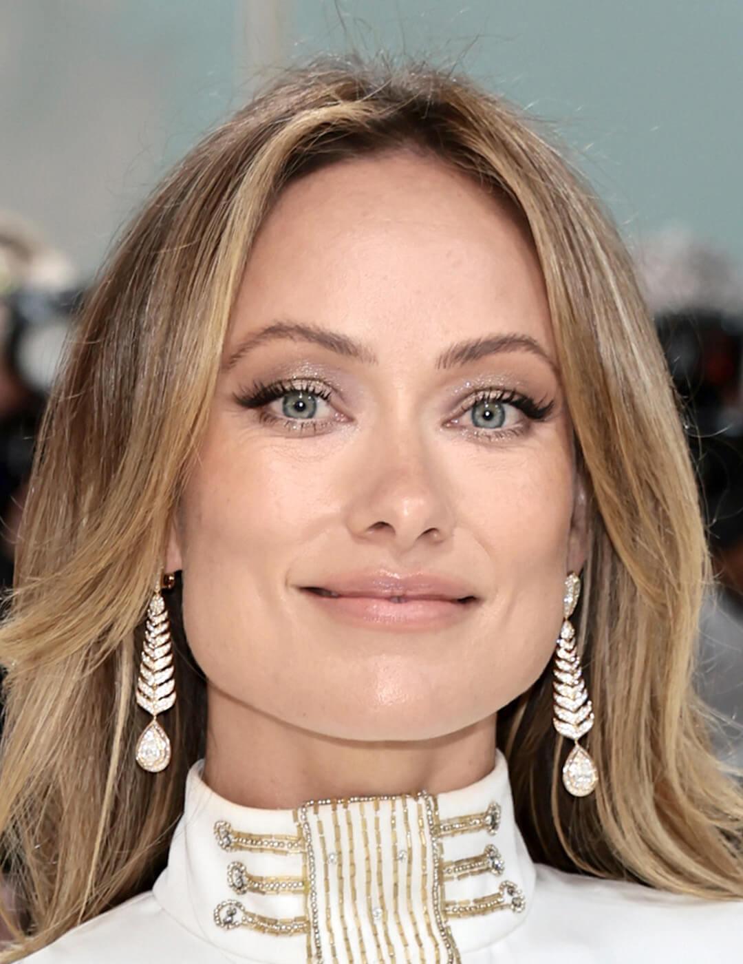 Close-up look of Olivia Wilde sporting a subtle makeup look, her blonde locks framing her face, and accessorizing with drop earrings, all while rocking a stunning white gown Mobile
