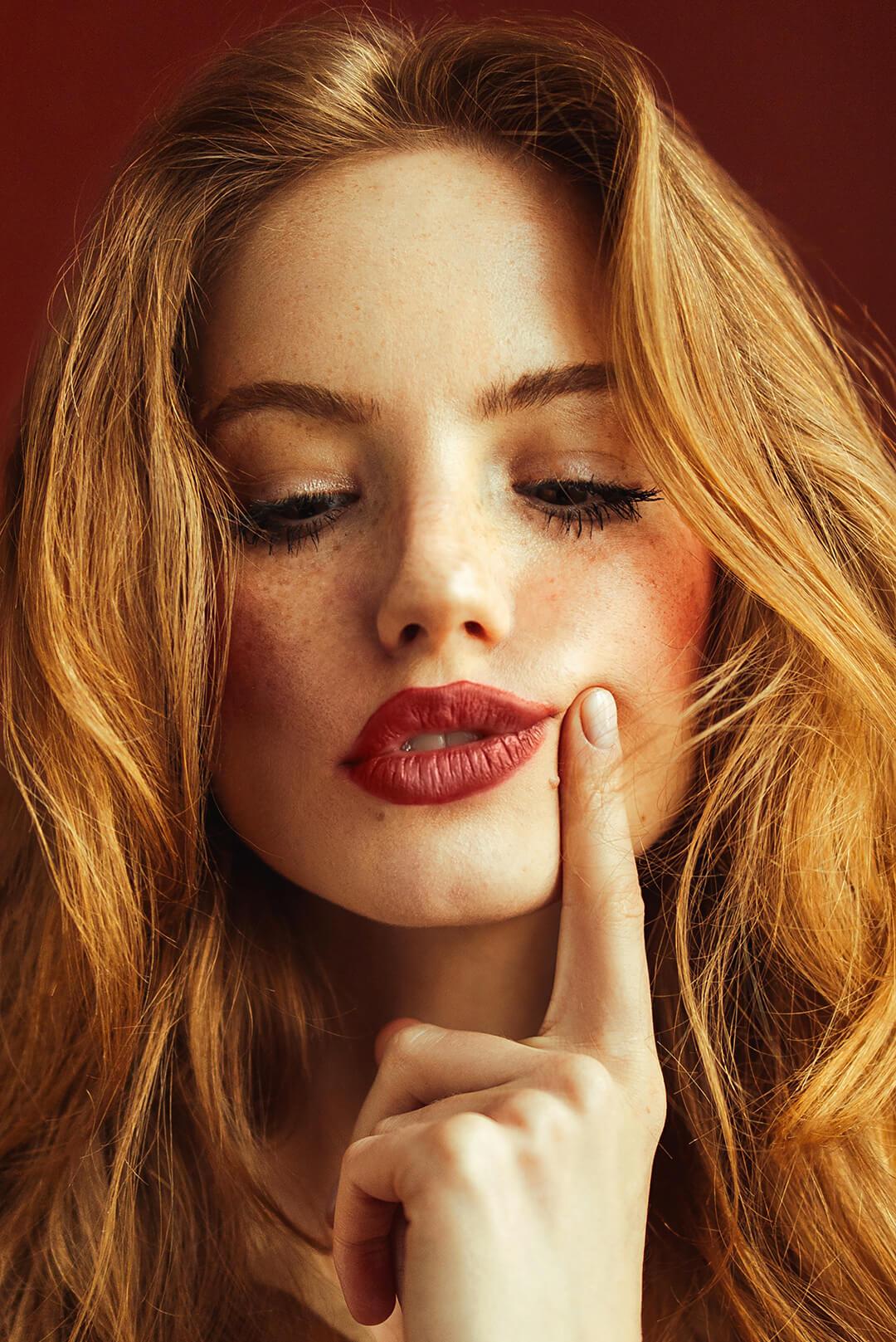 A photo of a woman with red hair and nude makeup look pressing her finger on cheeks Desktop