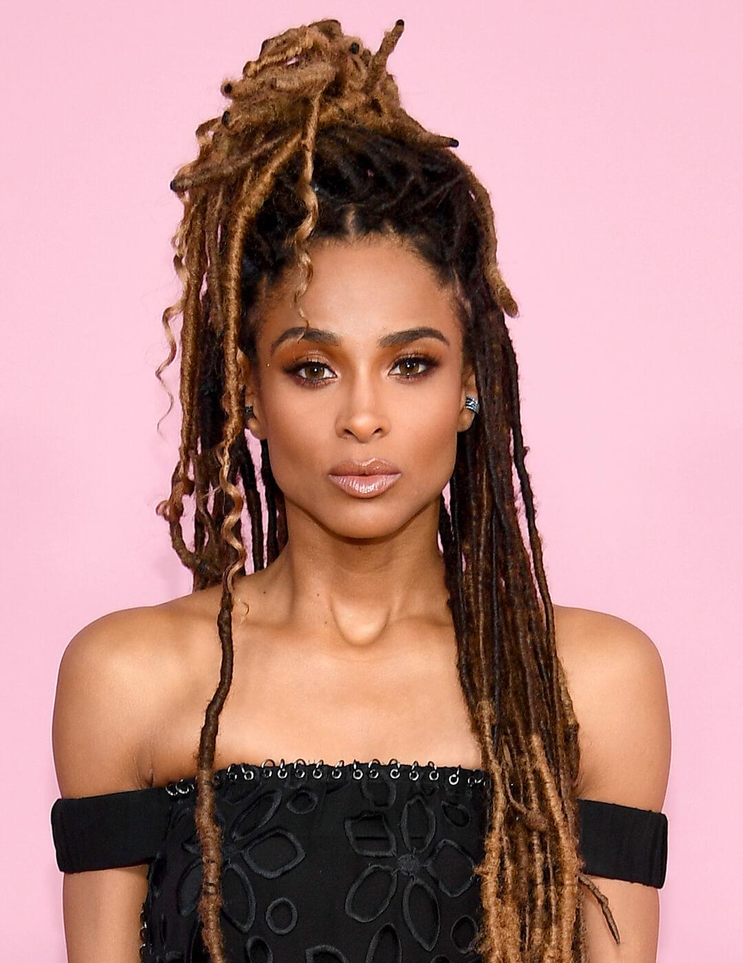 Close-up of Ciara rocking a faux locs half updo hairstyle, smoky eye makeup paired with a nude lipstick, and black dress against pink background Mobile