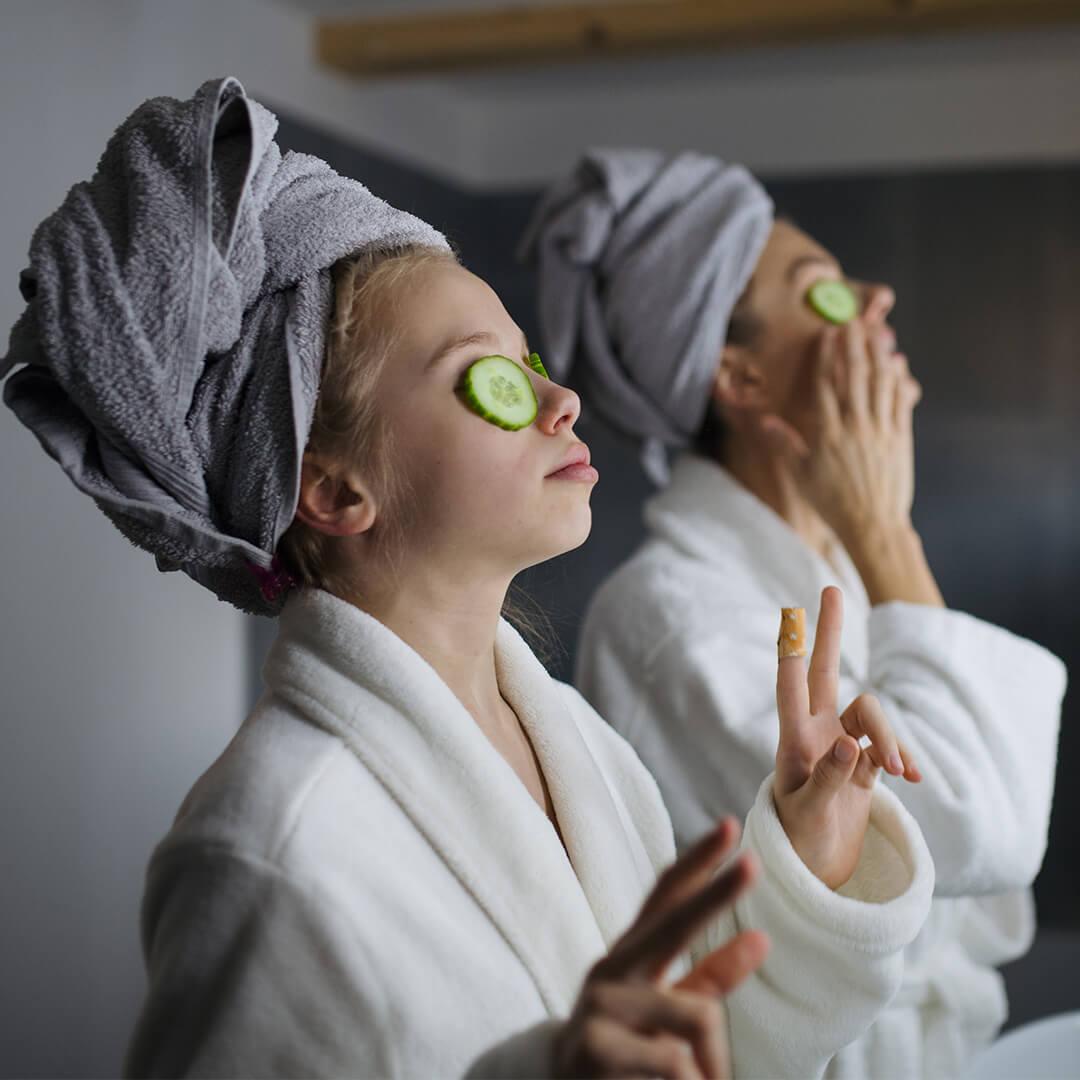 Mother and small daughter indoors in bathroom at home, putting cucumbers on eyes Desktop