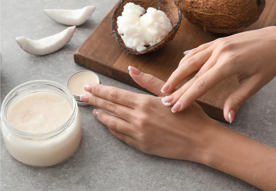 Close-up image of a woman applying hardened coconut oil onto skin and coconut husks on a chopping board on a grey surface Mobile