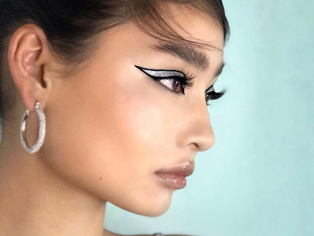 An image of a young woman striking a side-profile pose, elegantly adorned with black eyeliner and shimmering silver eyeshadow wearing silver hoop earrings and with a glossy lip finish Desktop