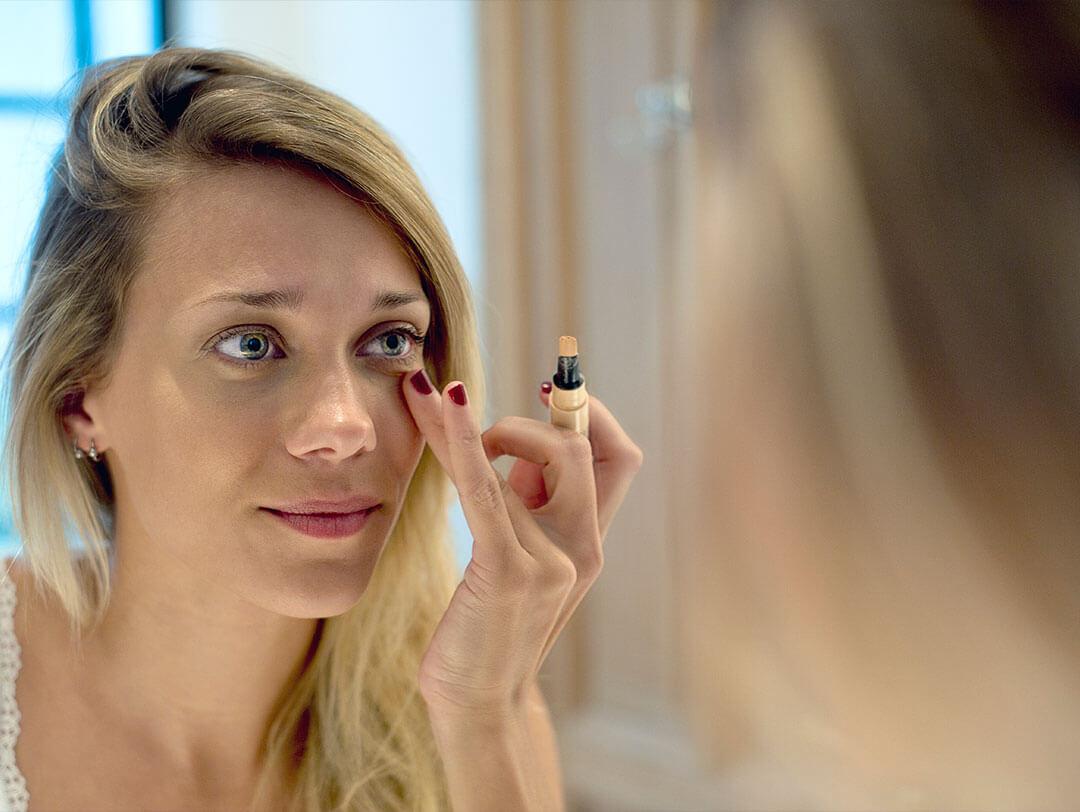 A woman using a concealer stick to apply makeup under her eyes, demonstrating techniques for hiding scars with makeup Desktop