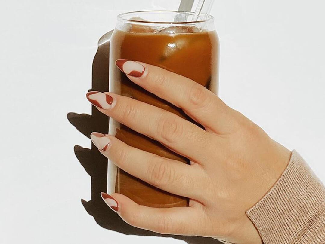 An image of a hand holding a cup of cold-brew coffee with a coffee-inspired nails on an offwhite background Desktop
