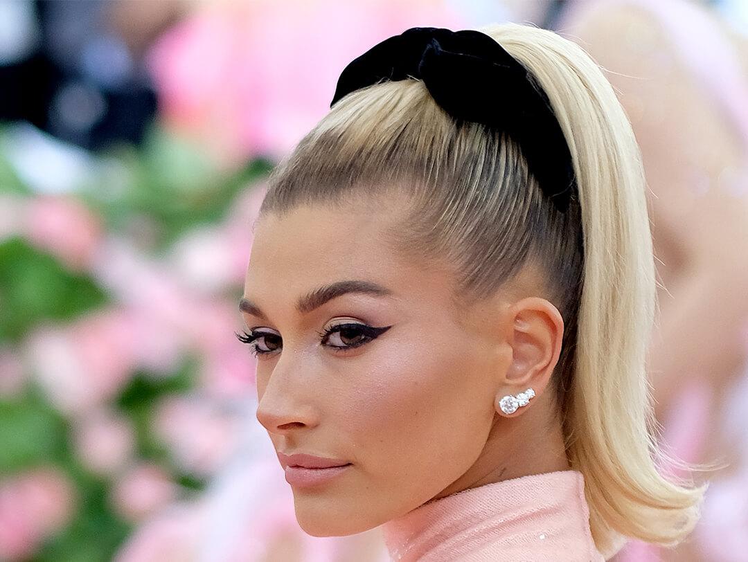 Close-up of Hailey Bieber in a high ponytail hairstyle, black headband, diamond earrings, pink dress, and bold cateye makeup look Desktop