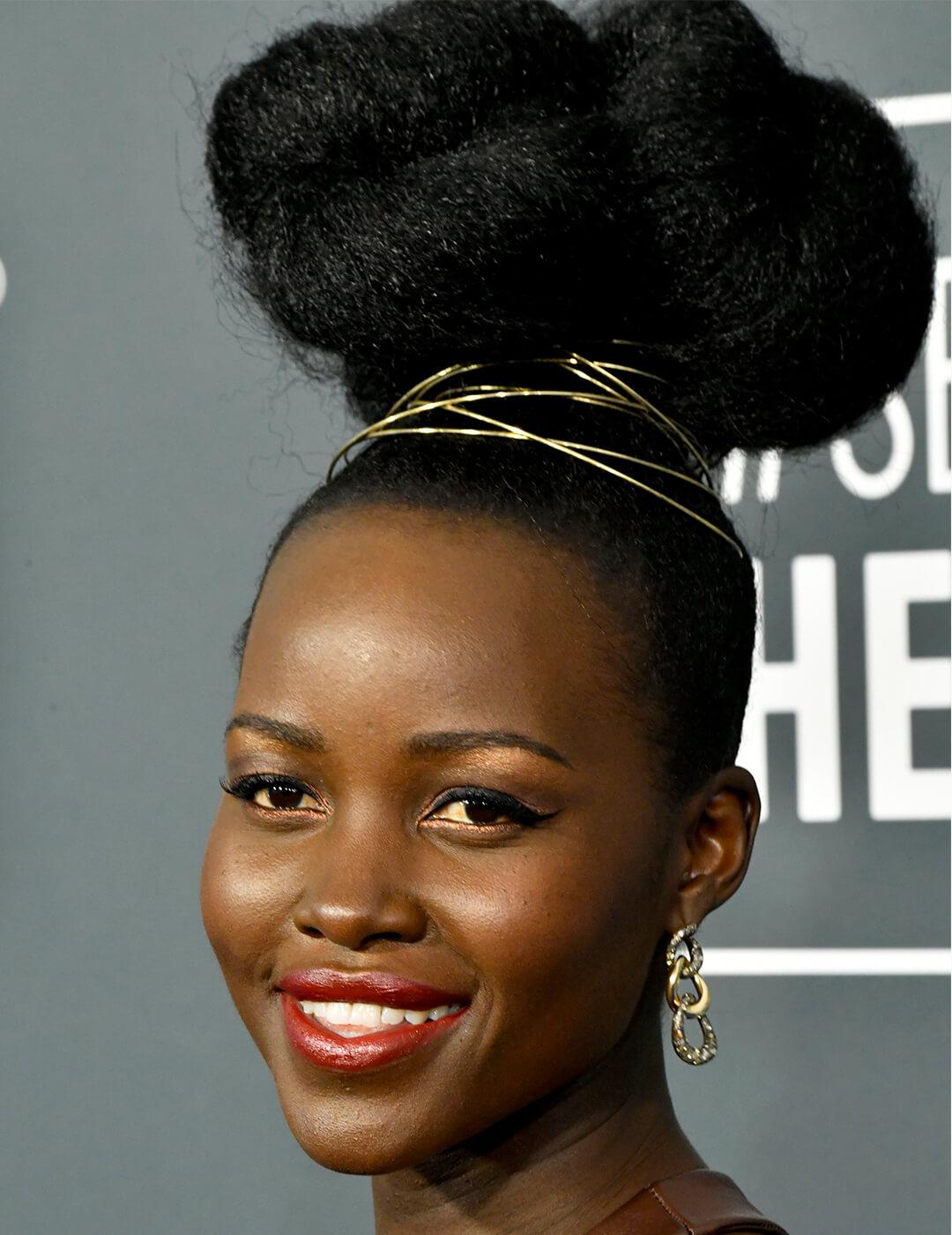 Close-up of Lupita Nyong'o rocking a high bun hairstyle wrapped with gold twine, copper eyeshadow makeup look paired with a berry lipstick, and gold links earrings Mobile