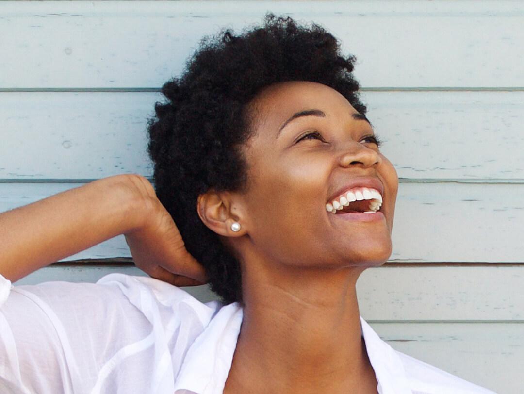 Side profile image of a black woman with short, curly hair in a white top smiling and posing against gray, wooden wall background Desktop
