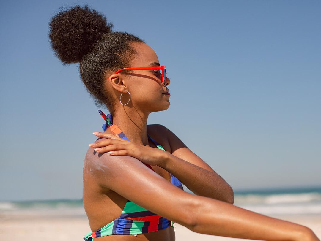 Woman of color in a colorful bikini, top bun, and red sunglasses putting on sunscreen on the beach Desktop
