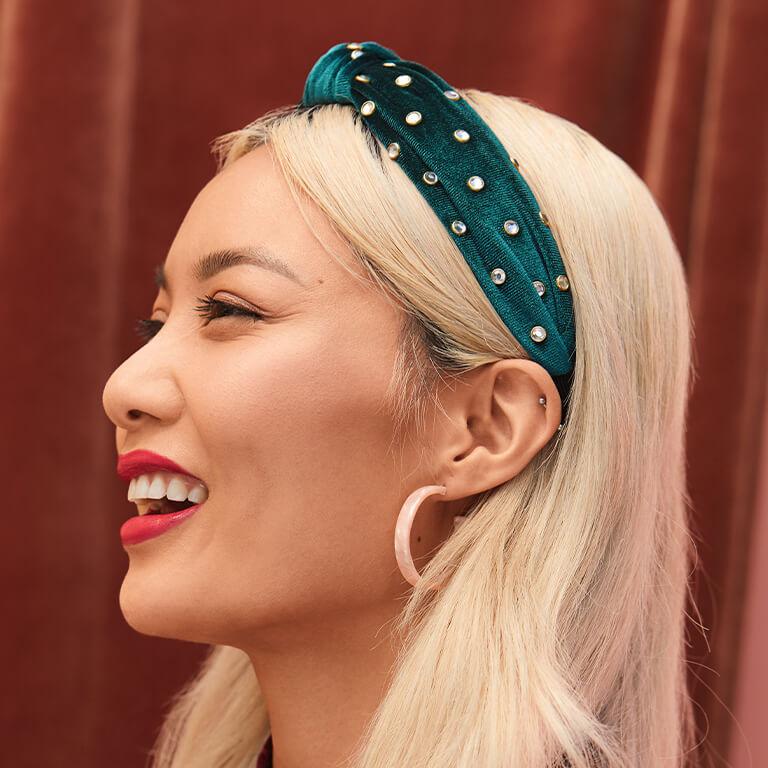 A profile image of a smiling model wearing gemstone-studded, green velvet headband and pink hoop earrings Desktop