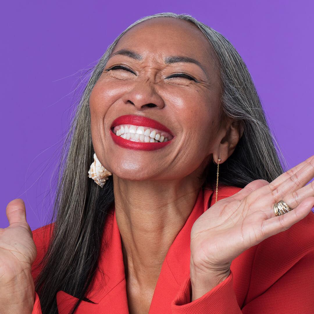 A photo of a mature woman laughing wearing red lipstick and sporting a red coat and conch earrings on a purple background Mobile