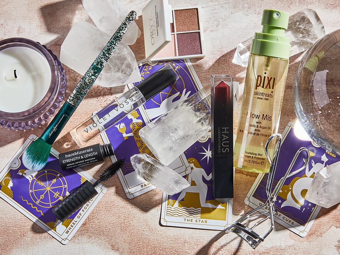 Flat lay image of skincare and makeup products and tools from various brands, tarot cards, crystals, and a white candle on a rose gold background Desktop