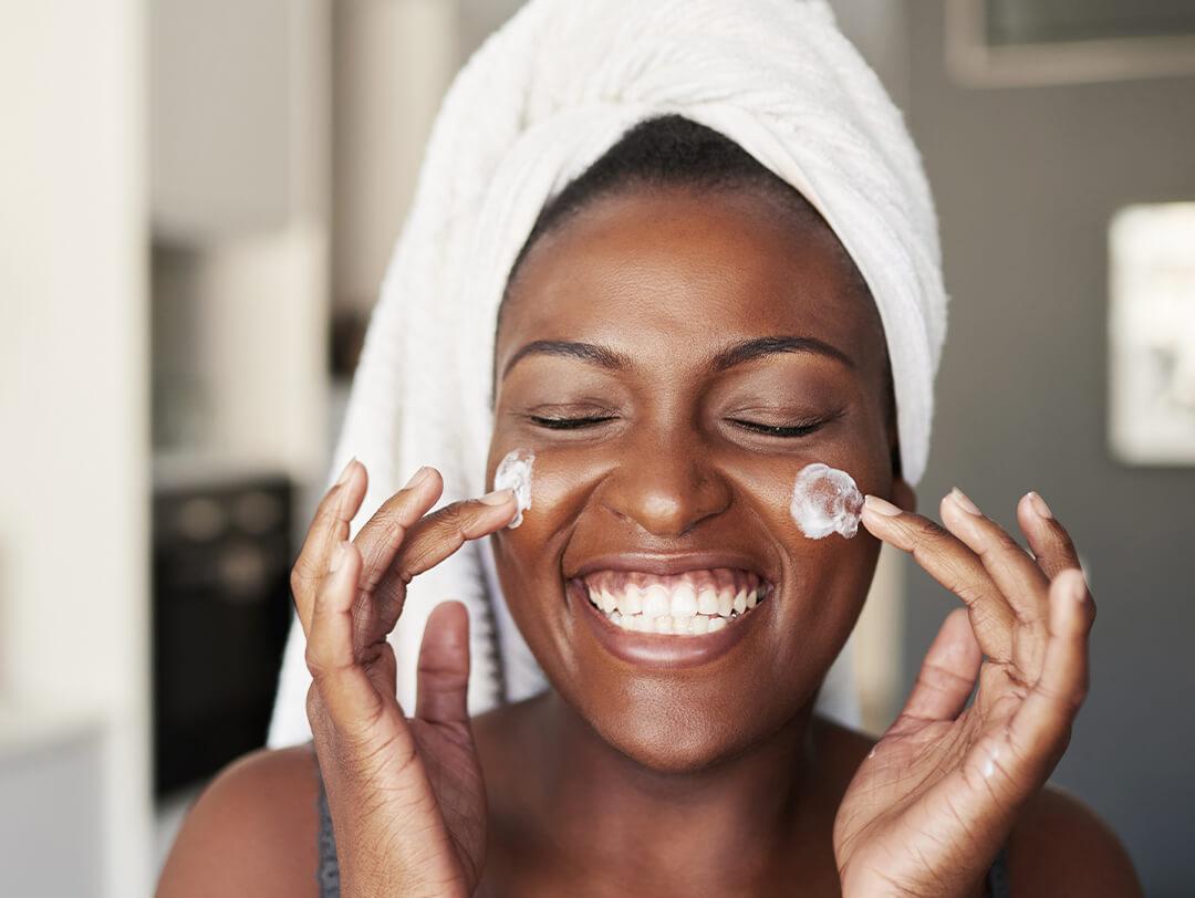 Image of a smiling Black woman with hair wrapped in white towel applying moisturizer on her cheeks Desktop