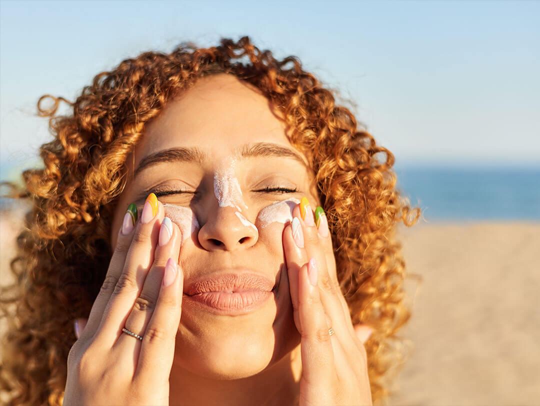 A close-up image of a young woman with curly blonde hair applying sunscreen to her face, her nails painted in bright colors Desktop