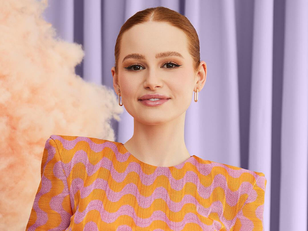Portrait of Madelaine Petsch looking glam in a wiggly orange and pink designed dress against lavender curtains Desktop
