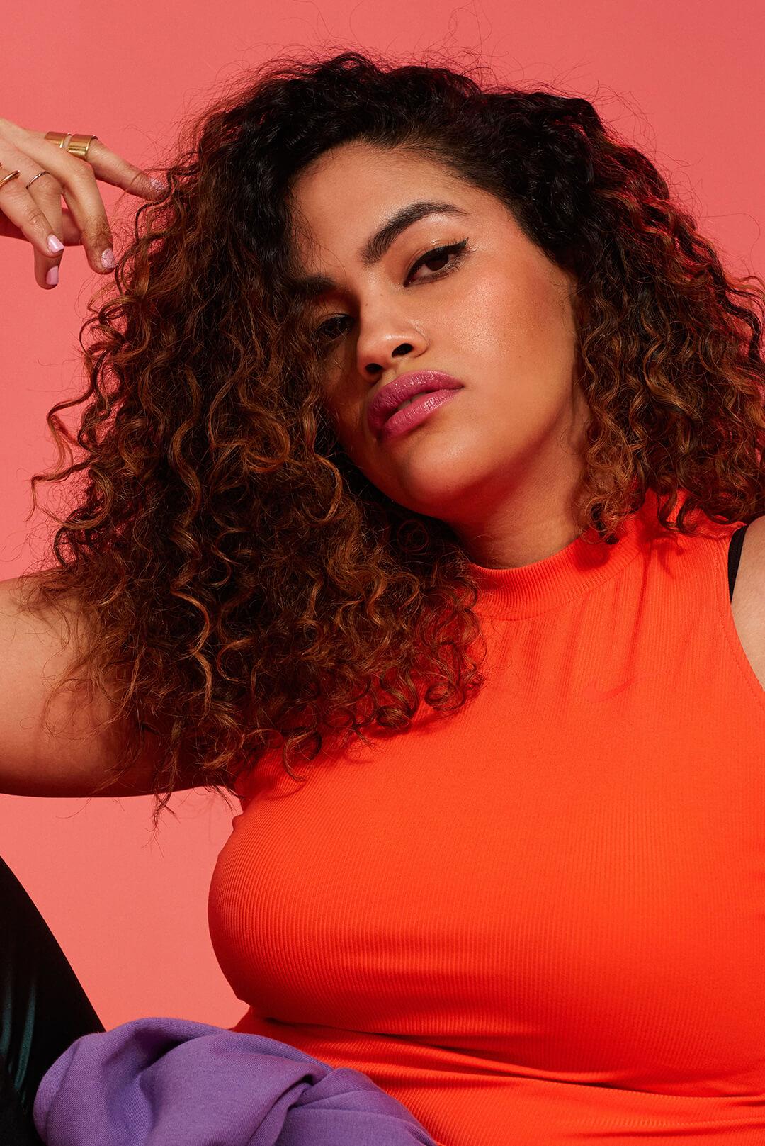 Close-up image of a model with thick, curly hair rocking a natural makeup look wearing an orange top on a red background Desktop