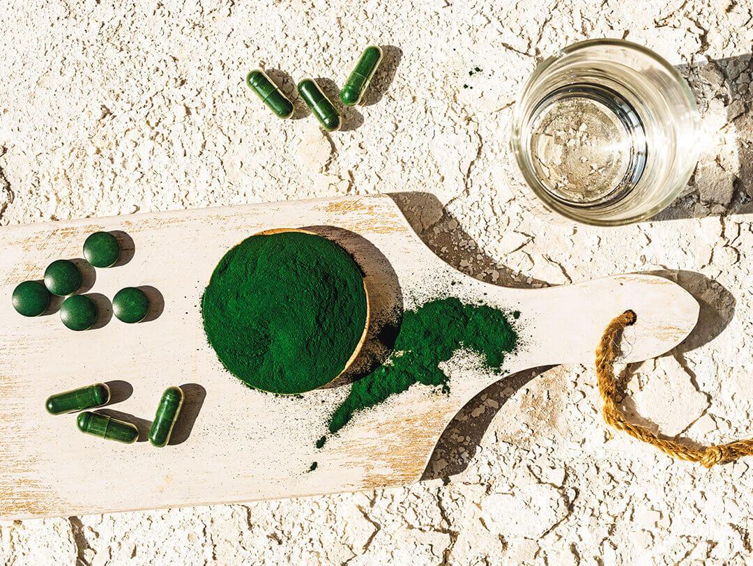 An image of a cup full of algae powder on a wooden board, pill capsules, and an empty jar Desktop