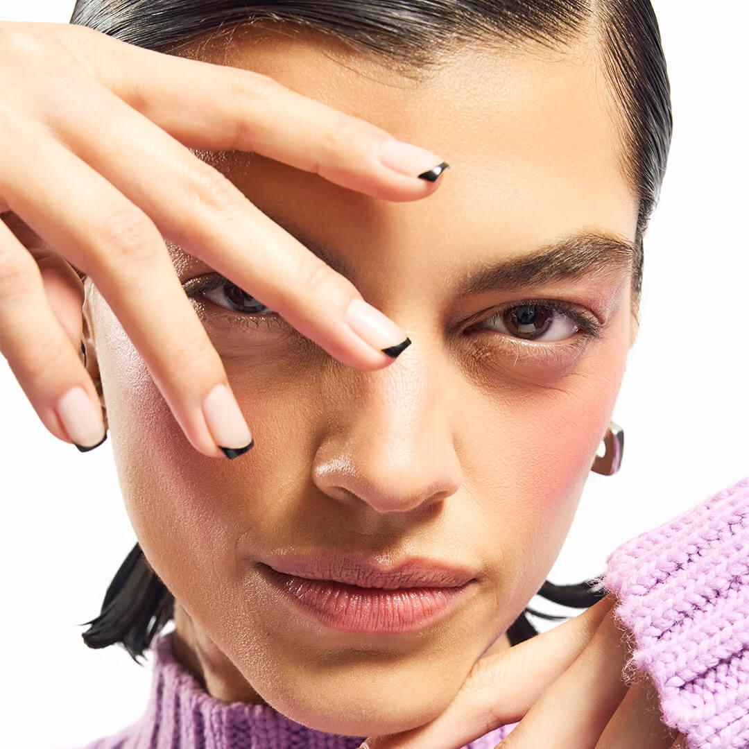 A close-up of a chic woman, her hand near her eye, highlighting her neatly shaped nude nails with sleek black tips Mobile
