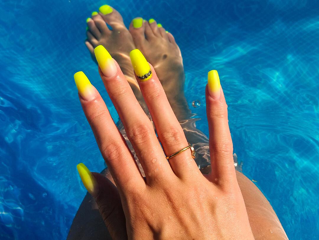 Close-up image of a woman's hand with long, neon yellow-colored nails against a pool background Desktop