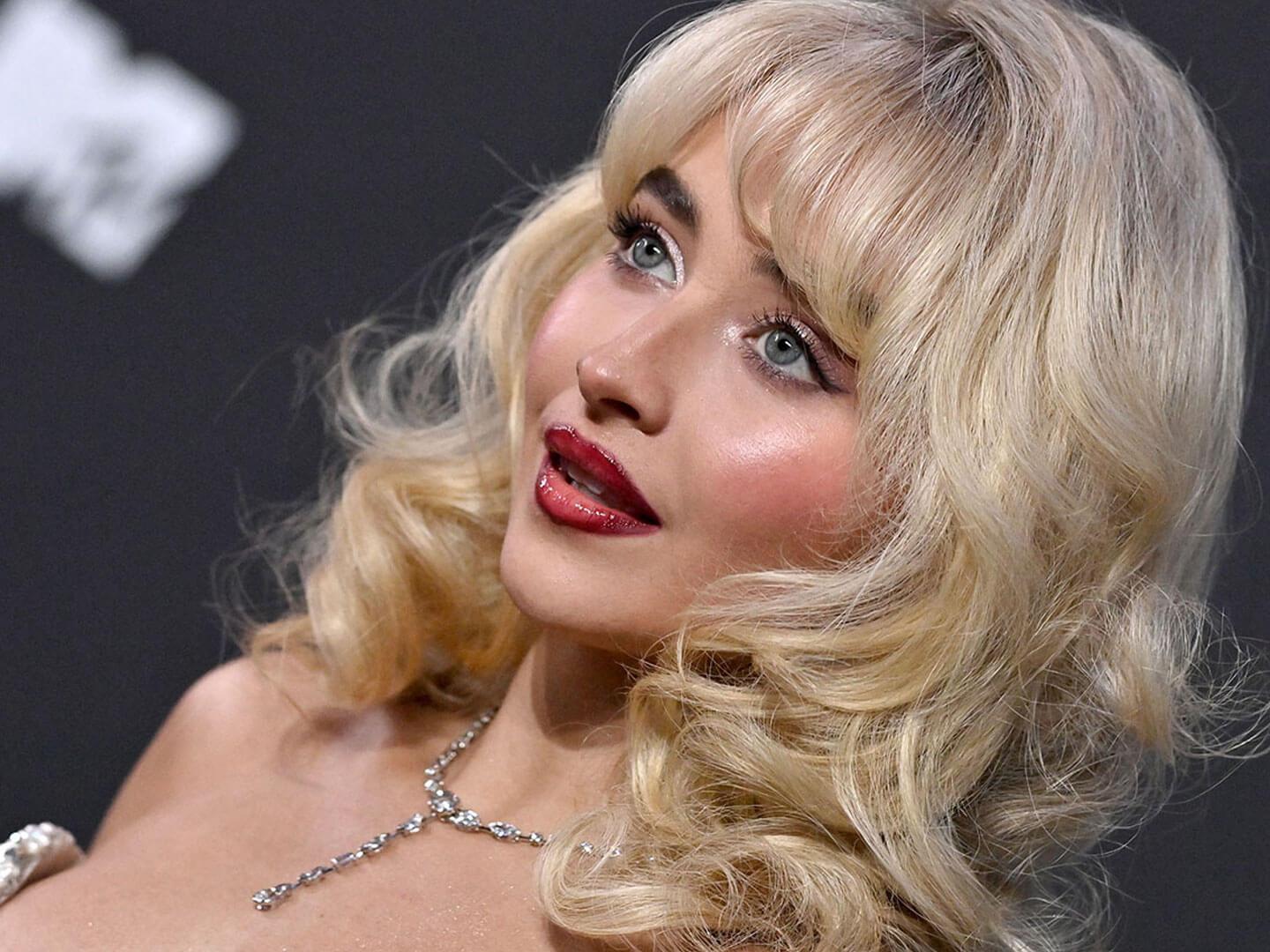 A blonde woman with glossy red lips, dramatic eyeliner, and soft curls, wearing a sparkling necklace, against a dark background Desktop