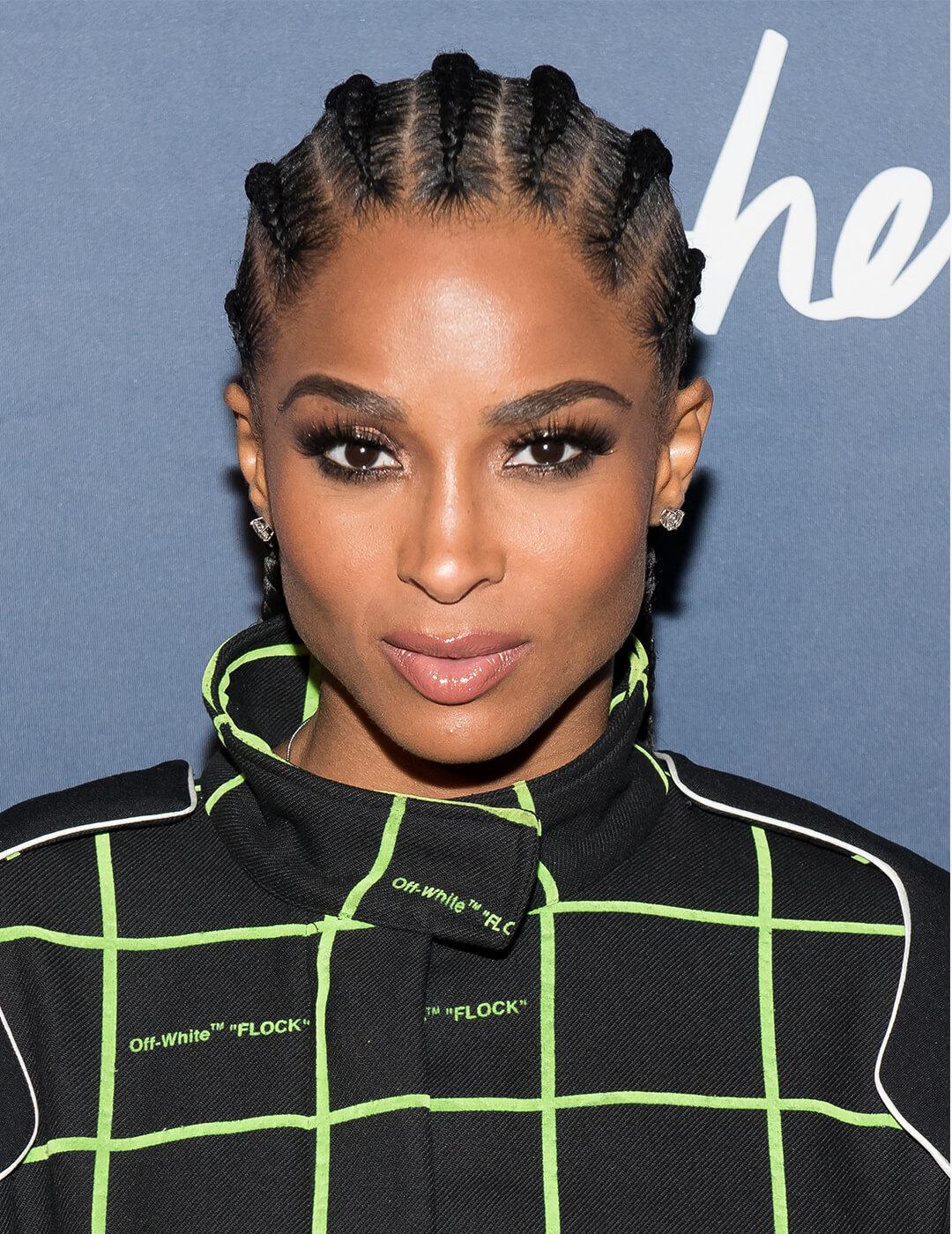 Close-up of Ciara rocking a cornrows hairstyle, smoky eye eyeshadow makeup paired with nude lips, and a black jacket with neon green intercrossing lines Desktop