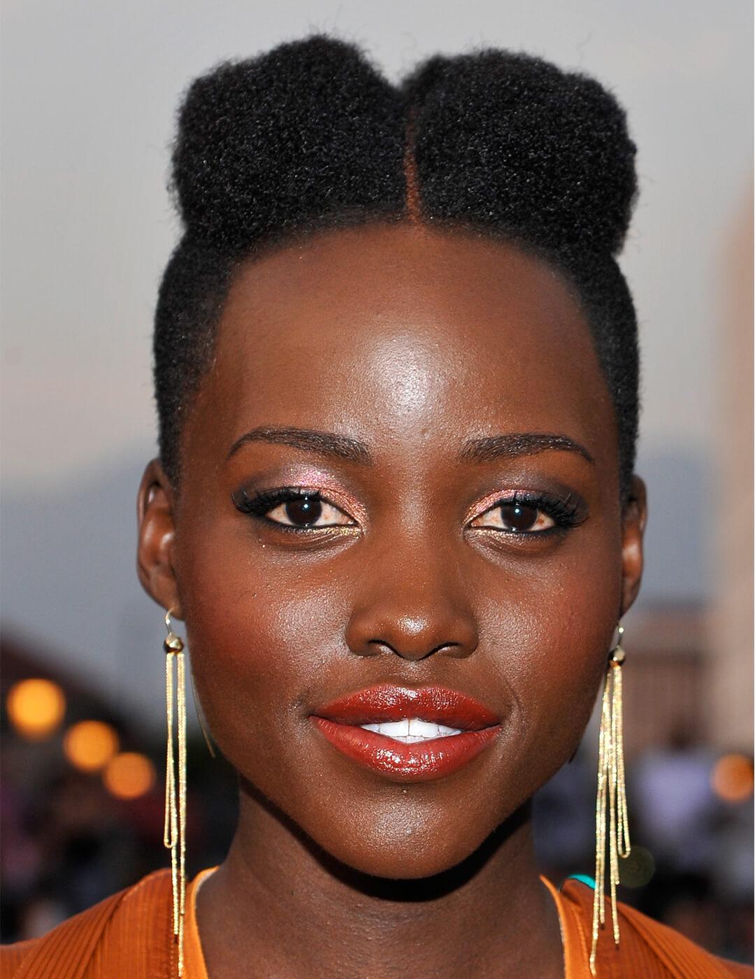 Close-up of Lupita Nyong'o wearing gold tassel earrings and orange dress rocking a faux mohawk hairstyle, shimmery coral eyeshadow and glossy coral lipstick makeup look Desktop