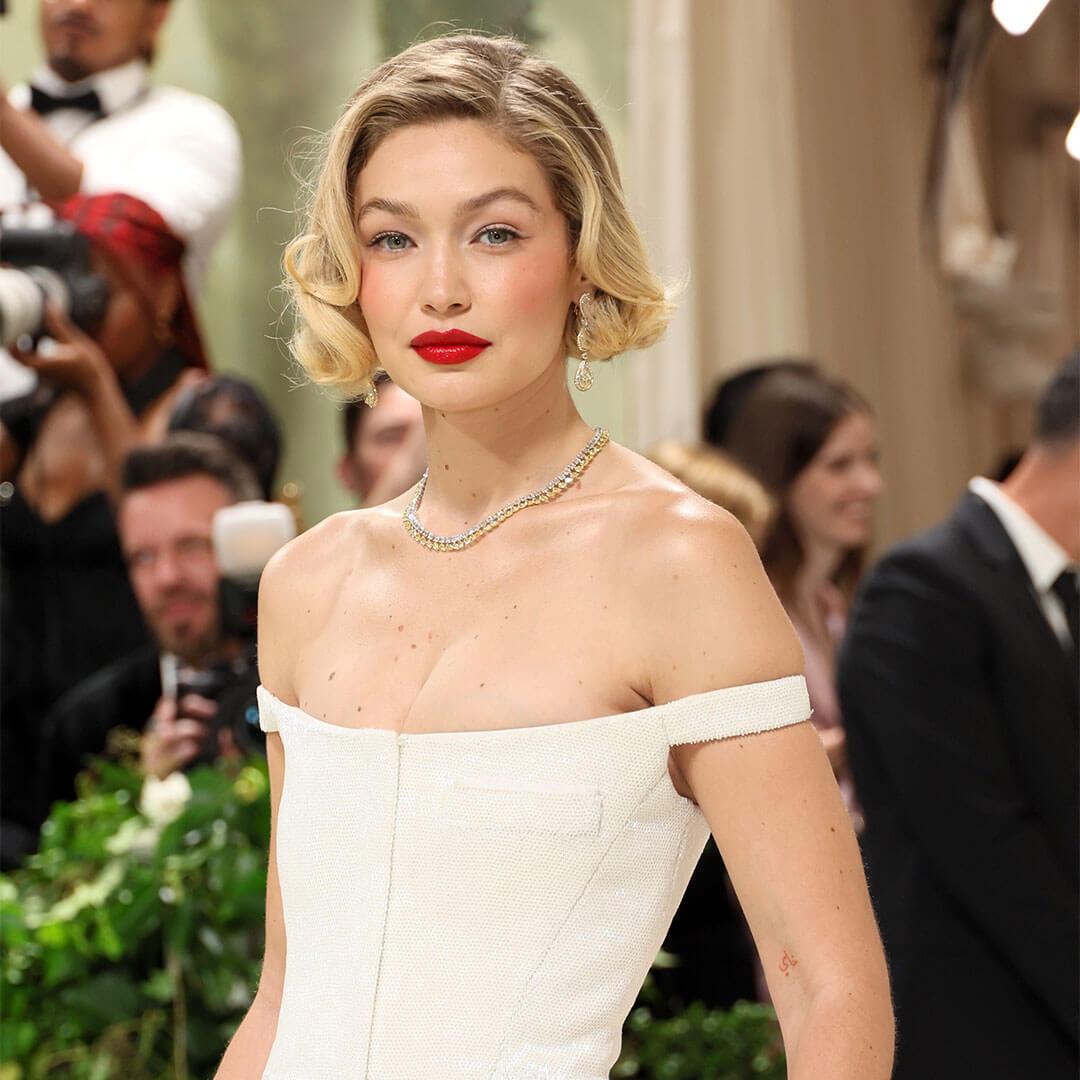 A photo of Gigi Hadid showing off her short blond hair, bright red lipstick, white off-the-shoulder dress, and diamond gold necklace with a monochromatic look Desktop