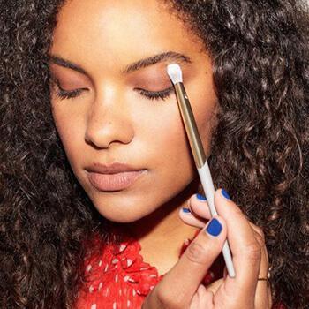 An image of a curly-haired model applying eye makeup on her crease using a COMPLEX CULTURE brush Mobile