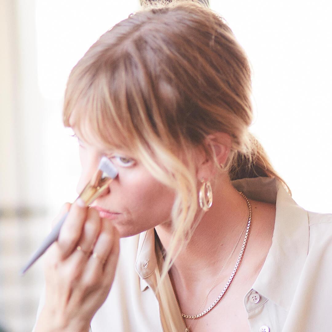 Model in a a cream button up blouse applying eye makeup under her eyes using a COMPLEX CULTURE makeup brush Mobile