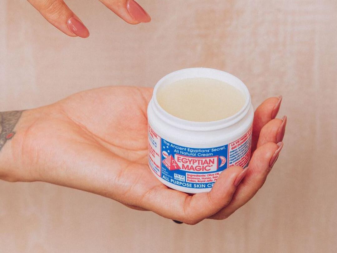 A closeup of a model's hand reaching for a EGYPTIAN MAGIC Multipurpose Cream on a linen background Desktop