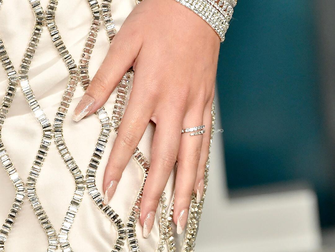 Close-up of Hailee Steinfeld's hand with silver bracelets and rings, and glittery French tip nail art resting on bejewelled cream dress Desktop
