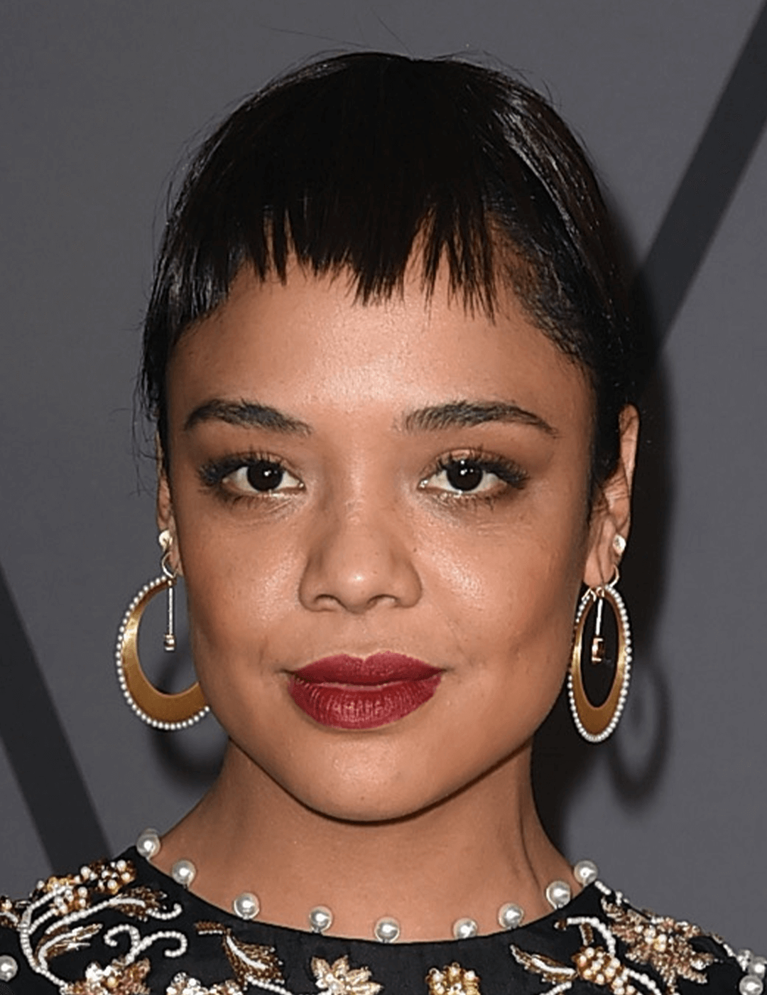 Tessa Thompson rocking a black and gold dress, neutral eye makeup with berry lips, gold hoop earrings, and a pixie cut hairstyle with micro bangs Desktop