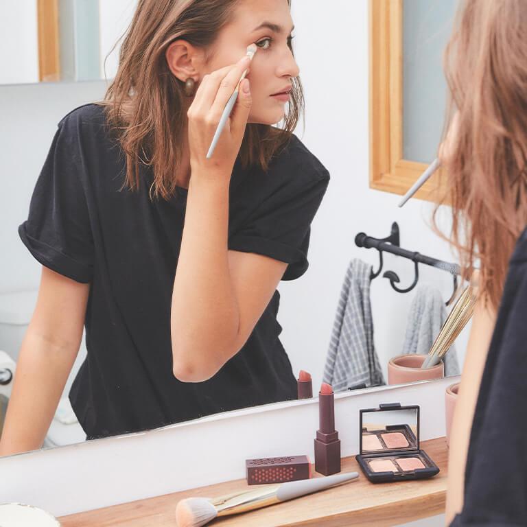 An image of a model in black shirt looking at herself in the mirror while using a COMPLEX CULTURE brush to apply eye makeup Desktop
