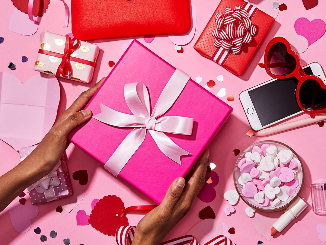 Image of a woman's hands holding a big pink gift box, small red and white gift boxes, heart-shaped confetti, bowl of heart-shaped candies, iPhone, red-framed heart sunglasses, and pink pen scattered on pink background Desktop