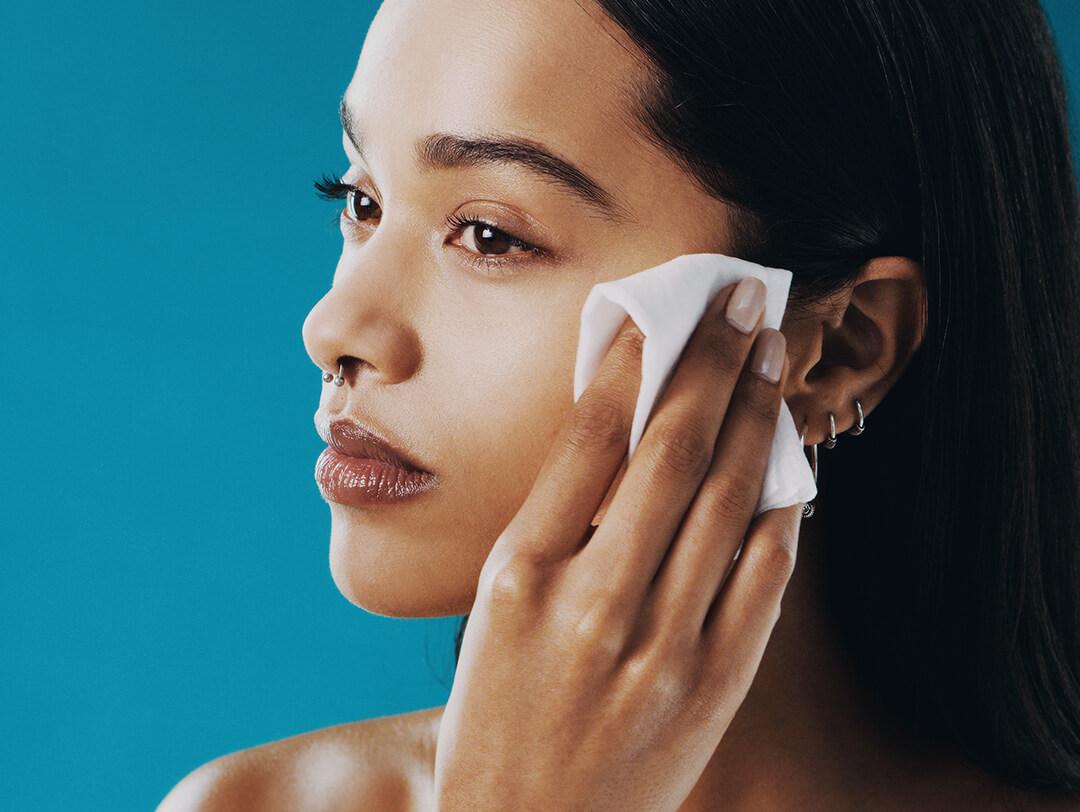 A photo of a model using a makeup remover wipe to clean her face from makeup on a blue-green background Desktop
