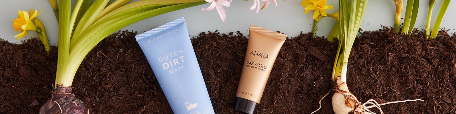 A flatlay image of BLOOMEFFECTS Dutch Dirt Mask and AHAVA 24K Gold Mineral Mud Mask and orchids on soil dirt Desktop