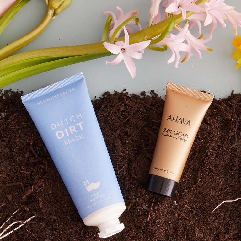 A flatlay image of BLOOMEFFECTS Dutch Dirt Mask and AHAVA 24K Gold Mineral Mud Mask and orchids on soil dirt Mobile
