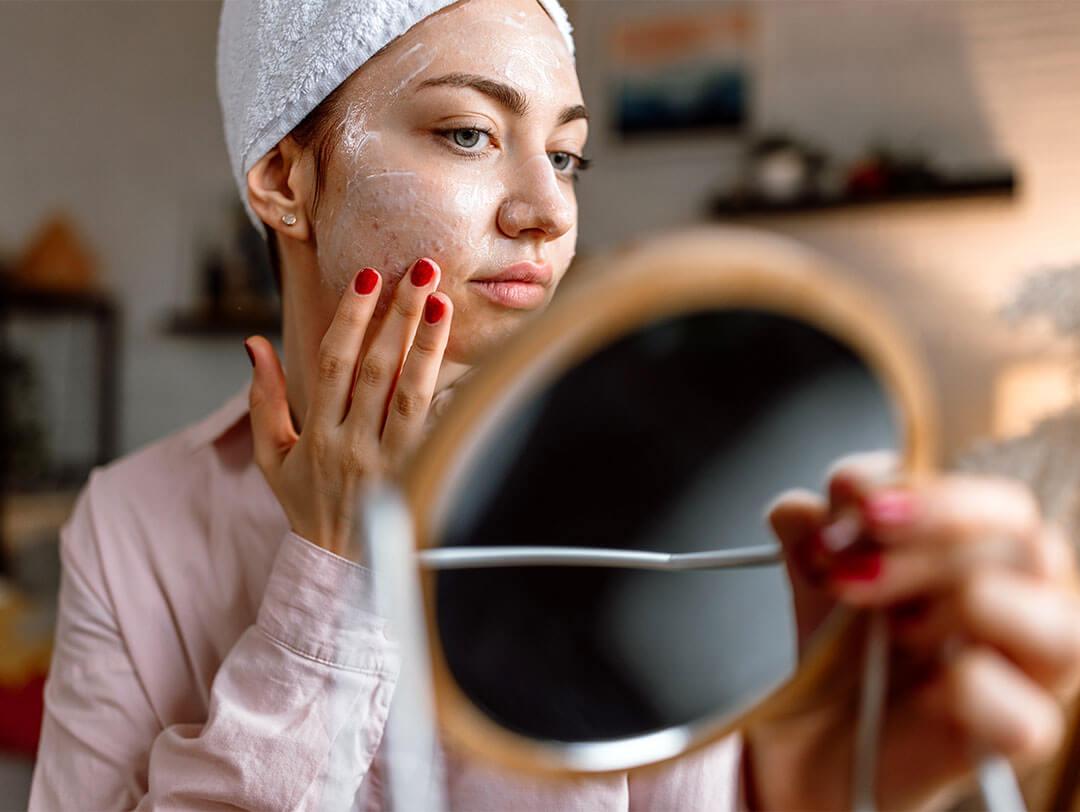 An image of a young woman with acne skin applying cream on her face while holding a mirror Desktop