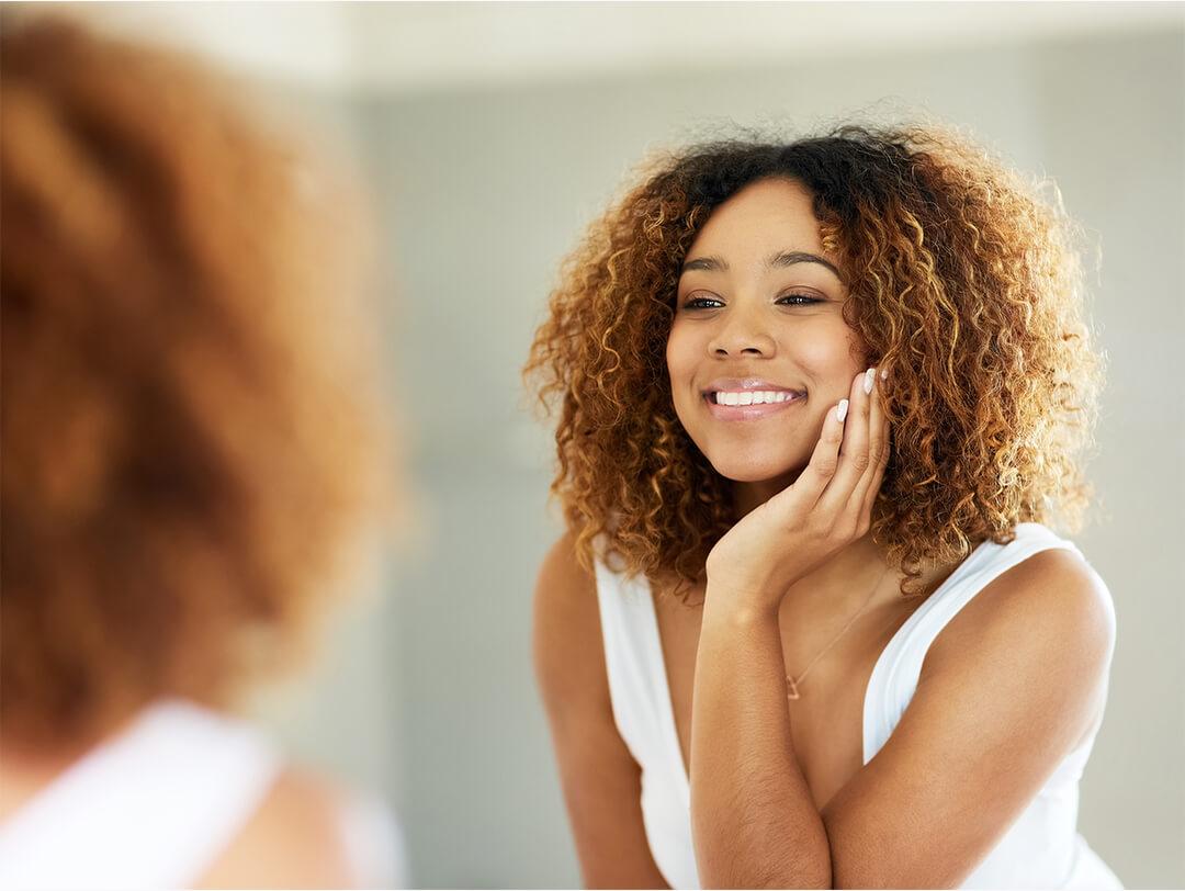 Close-up of a Black woman with curly hair touching her face while looking at herself in the mirror Desktop