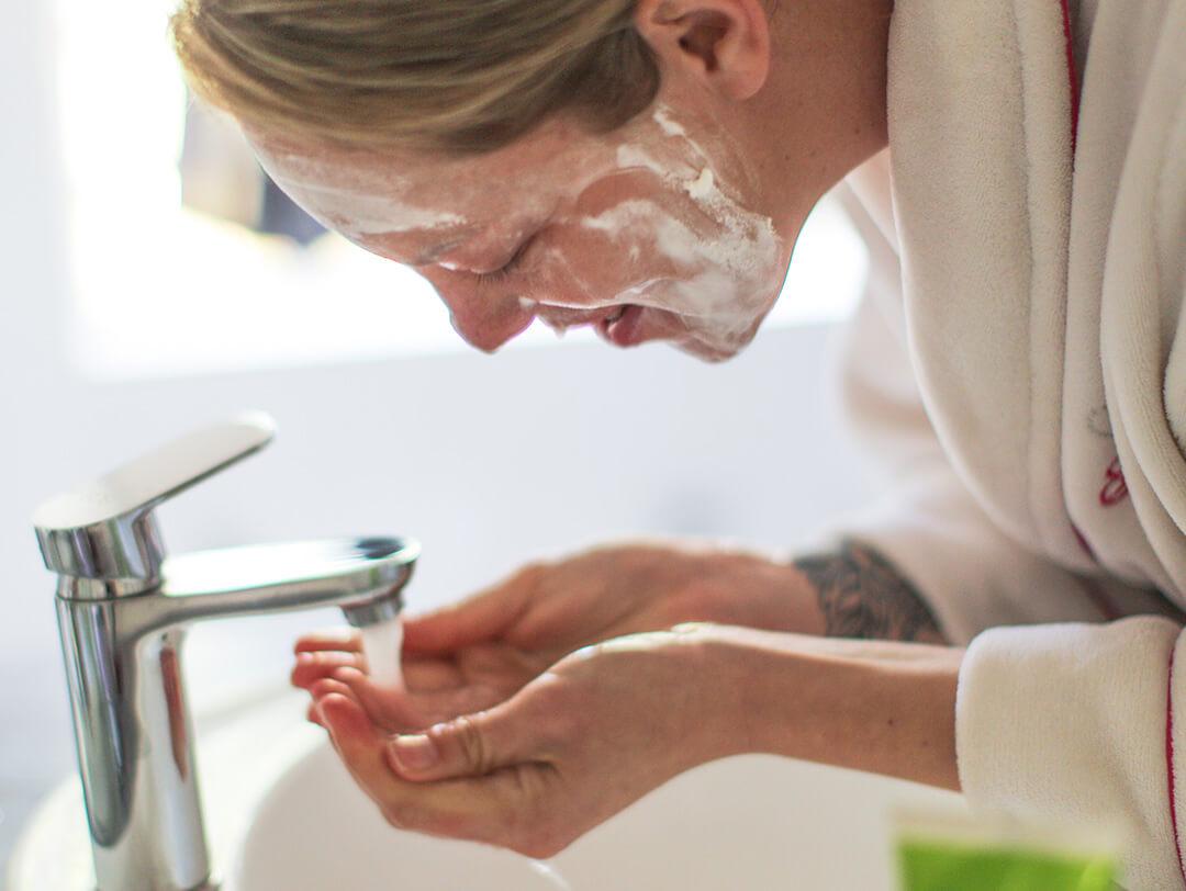 A woman wearing a white robe washing her face in the sink while wearing a facial mask Desktop
