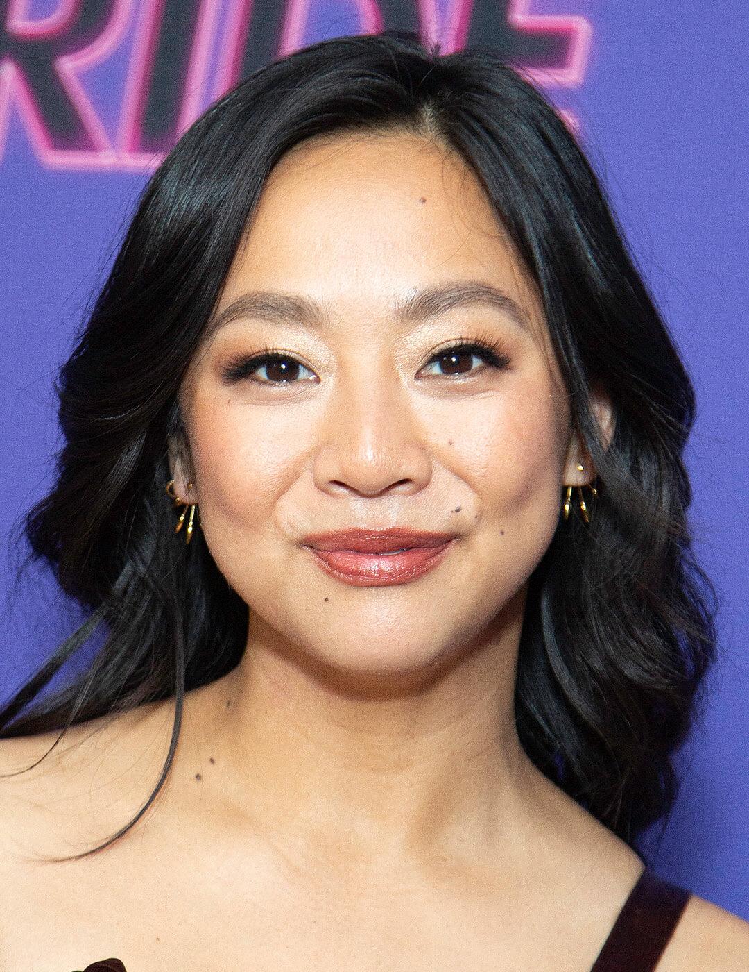 Stephanie Hsu smiling at the camera wearing a black gown against a purple background Mobile
