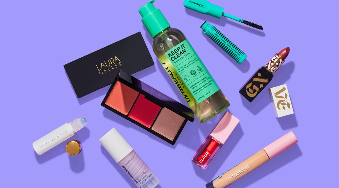 Skincare, makeup, and hair care products and tools from various brands scattered on purple background Desktop