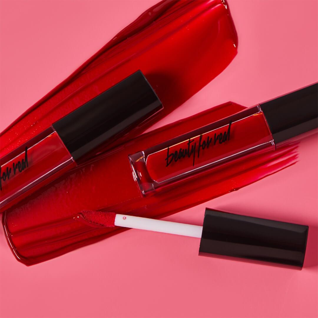 Image of two tubes of BEAUTY FOR REAL Lip Cream + Color in Really Red with applicator swatched on dark pink background Desktop