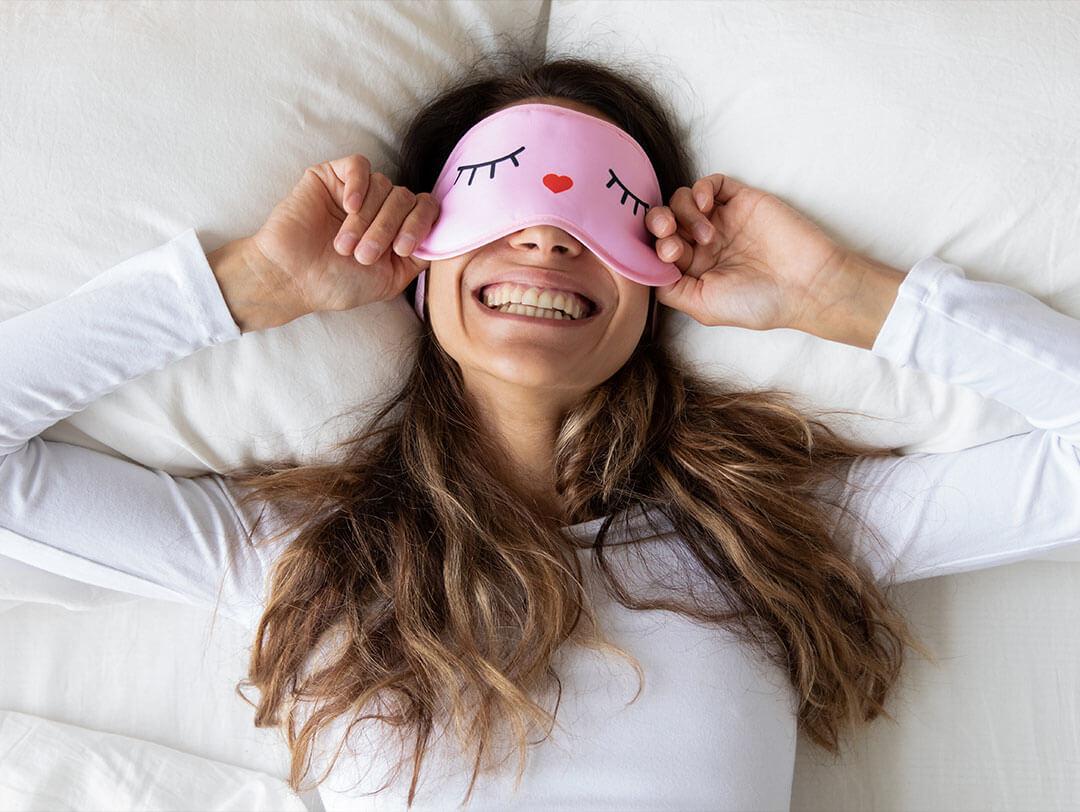 An image of a smiling woman with a cute pink sleeping mask is nestled in bed, wearing a cozy white long-sleeve shirt Desktop