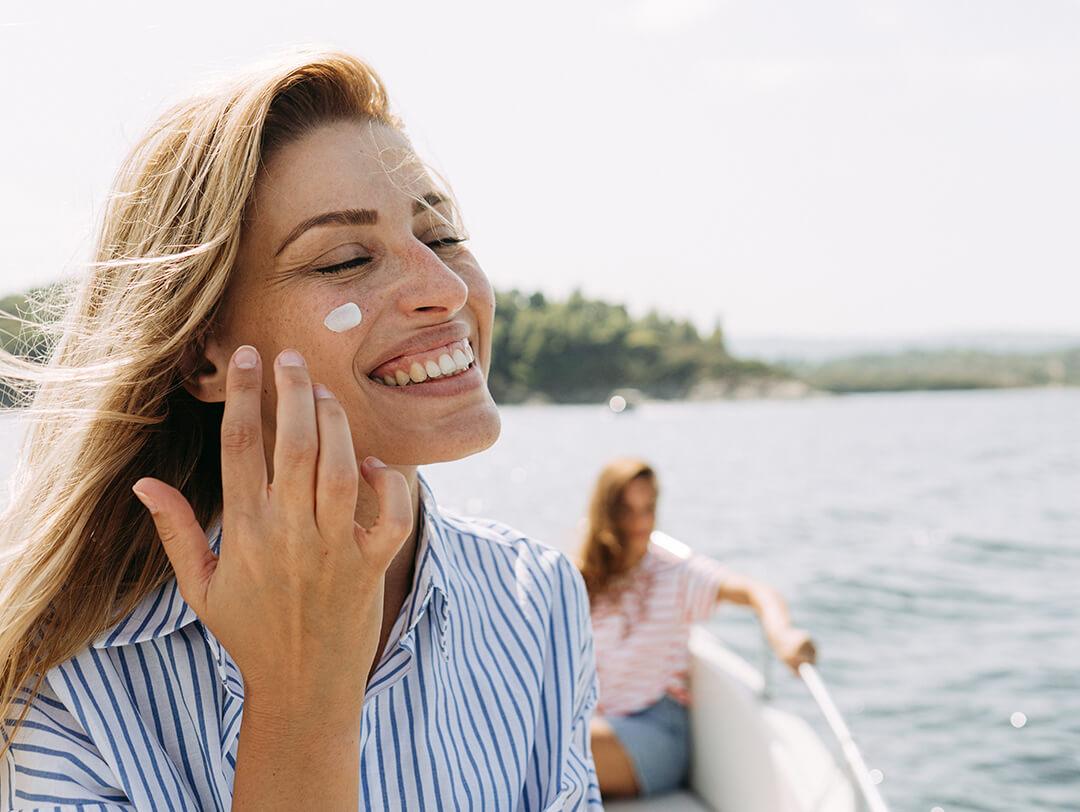 Photo of a smiling woman applying sunscreen while taking a boat ride with her friend Desktop