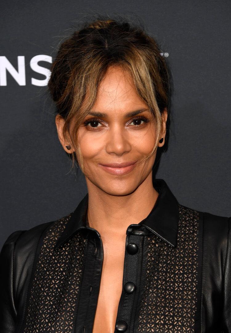 A close-up photo of Halle Berry wearing an open-buttoned black leather one-piece suit boasting curtain bangs hairstyle donned a chic black earring Mobile