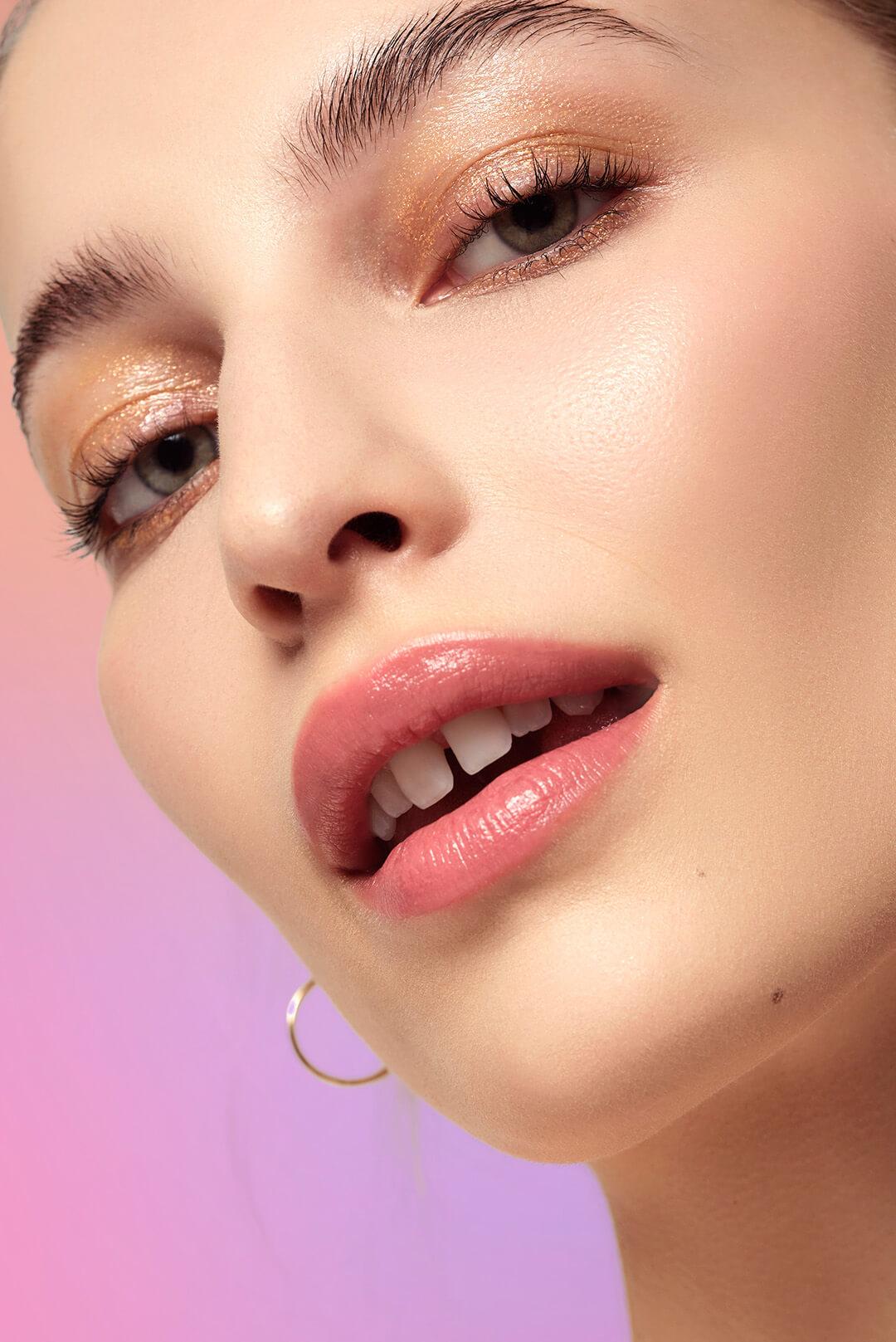 A closeup photo of a model with glossy makeup look on a pink gradient background Desktop