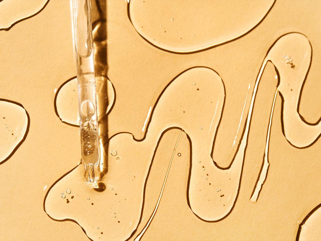 A picture of a dropper with serum drops splattered against a soft yellow backdrop Desktop