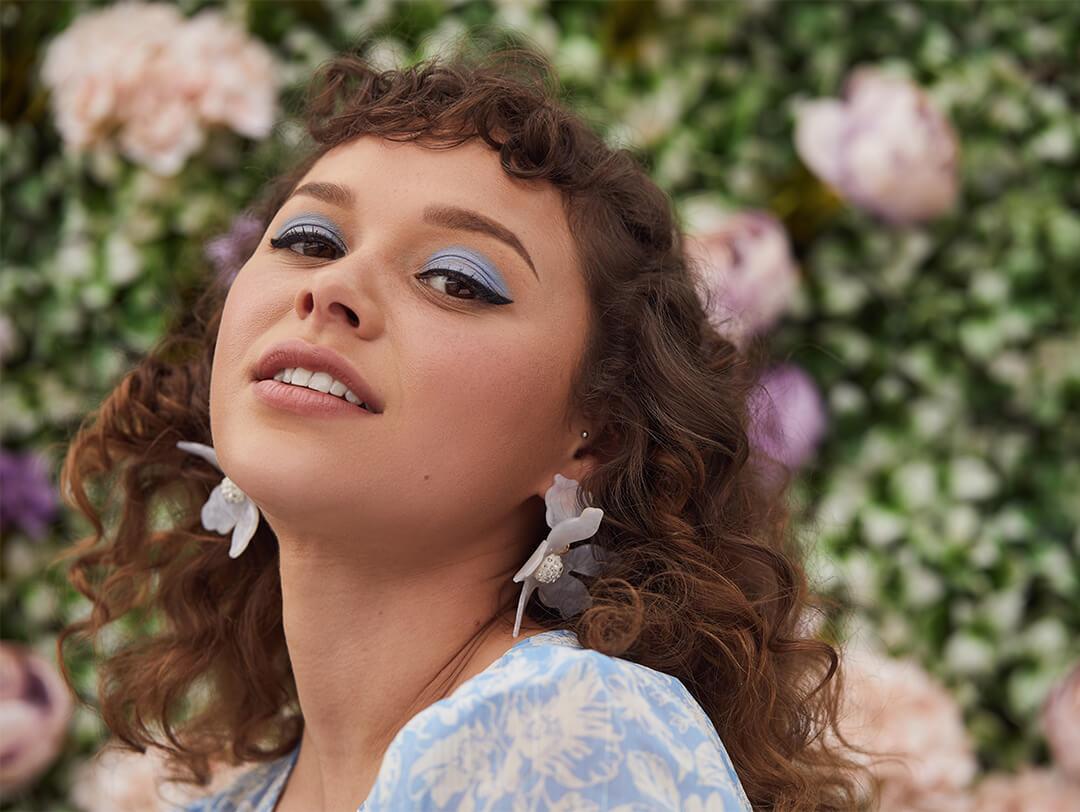 Close-up image of a mid-length curly haired model wearing blue eyeshadow, white flower earrings, and blue and white floral outfit posing in a garden Desktop