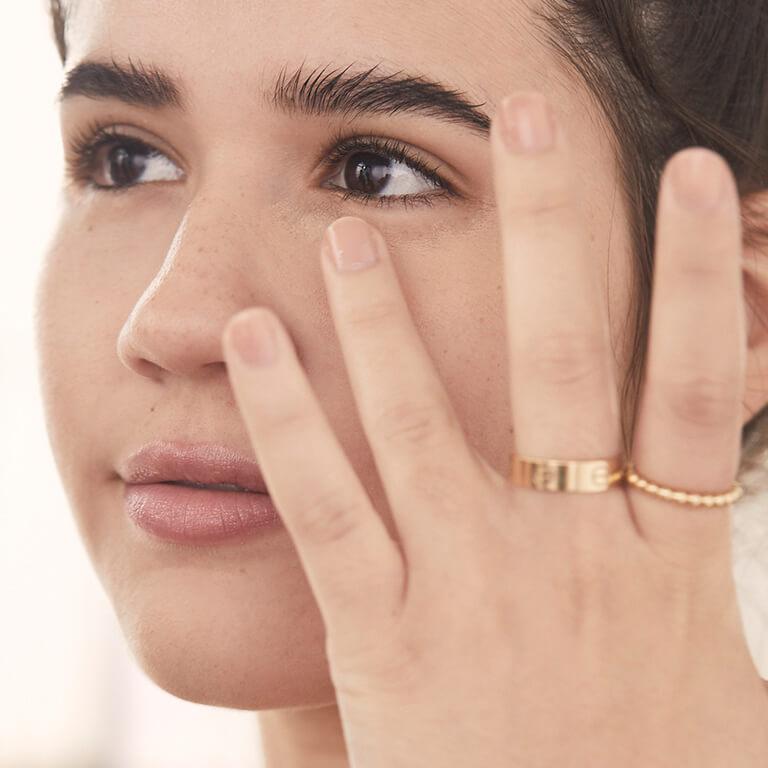 A closeup image of a model wearing gold rings applying concealer under eyes with her ring finger Desktop