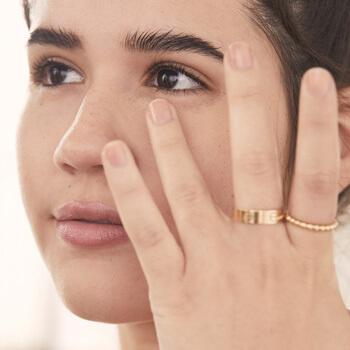 A closeup image of a model wearing gold rings applying concealer under eyes with her ring finger Mobile